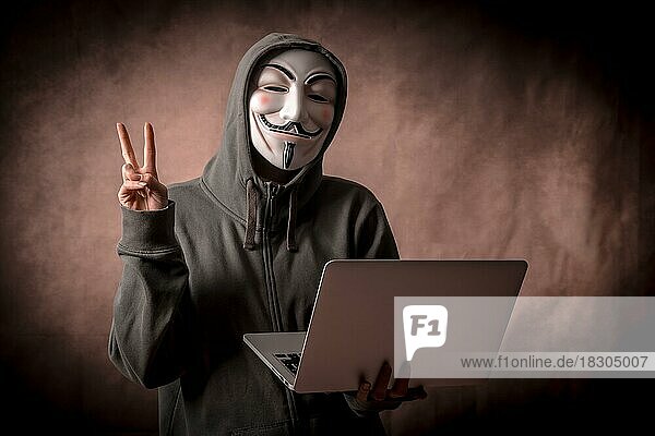 Hacker mit anonymer Maske mit einem Laptop  der das Siegeszeichen macht  Studioaufnahme