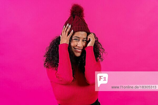 Lockenköpfige Frau mit Wollmütze vor rosa Hintergrund  schüchtern lächelnd  Studioaufnahme