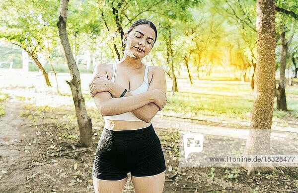 Junge Frau in Sportkleidung umarmt sich glücklich und positiv in einem Park. Lächelnde Sportlerin  die sich umarmt und frische Luft atmet. Ein sportliches Mädchen atmet glücklich und voller Luft umarmt sich selbst