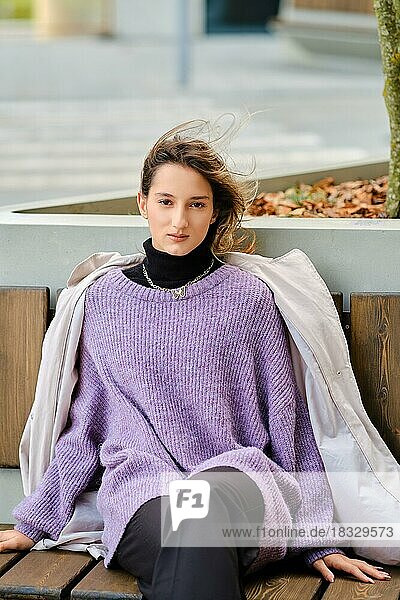Junge Frau in weiten Hosen  Pullover  Regenmantel und groben Stiefeln sitzt auf einer Bank und schaut geradeaus