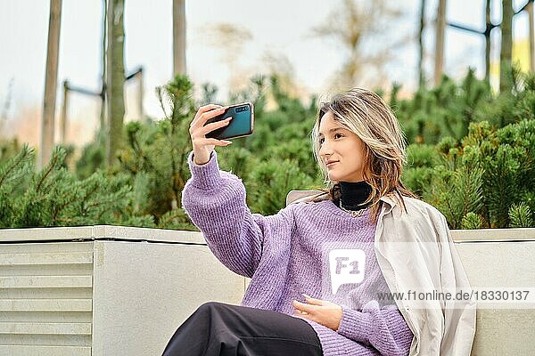Süße junge Frau macht Selfie oder Videoanruf  während sie auf einer Bank in der Stadt sitzt