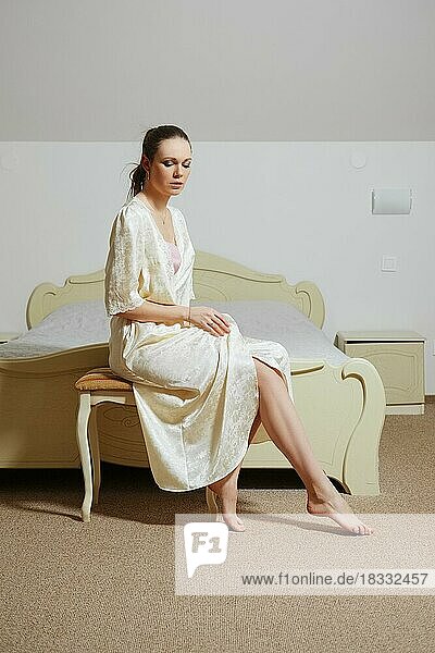 Hübsche junge Dame im Schlafzimmer in Seiden-Peignoir sitzt auf der Couch in der Nähe des Bettes