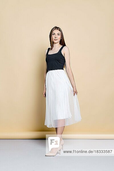 Full length portrait of pretty young woman in black tank top and white skirt posing over pale yellow background