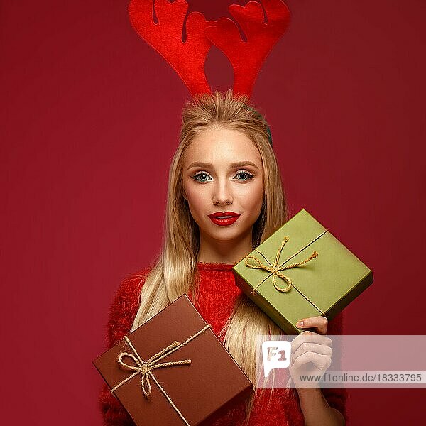Schönes blondes Mädchen in einem Neujahrsbild mit Kisten mit Geschenken in den Händen und Hirschhörnern auf dem Kopf. Schönes Gesicht mit festlichem Make-up. Foto im Studio aufgenommen
