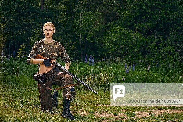 Ernstes Jägermädchen mit doppelläufigem Gewehr auf einem Knie stehend im morgendlichen Wald