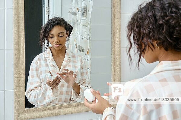 Attraktive afroamerikanische Frau schaut in den Spiegel und trägt Gesichtscreme im Badezimmer auf