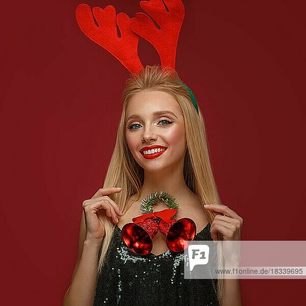 Schönes blondes Mädchen in einem Neujahrsbild mit Weihnachtsglocken um ihren Hals und Hirschhörnern auf ihrem Kopf. Schönes Gesicht mit festlichem Make-up. Foto im Studio aufgenommen