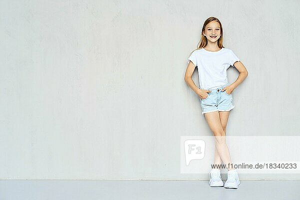 Junges sportliches Mädchen in weißem T-Shirt und Jeansshorts lehnt mit beiden Händen in den Taschen an der Wand