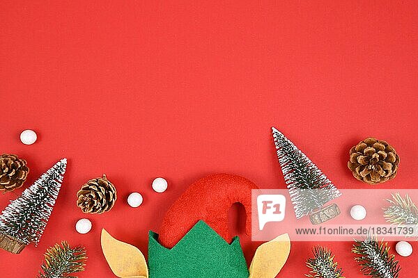 Saisonales Flatlay mit Weihnachtselfenmütze und -ohren  kleinen Tannenbäumen und -zapfen und weißen Schneebällen unten und leerem Kopierbereich auf rotem Hintergrund oben