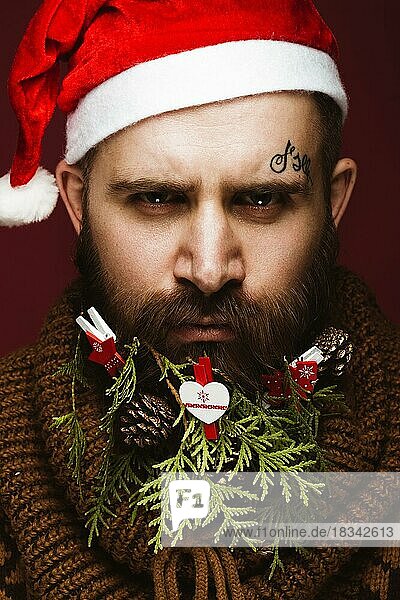Lustiger bärtiger Mann in einem Neujahrsbild als Weihnachtsmann mit Dekorationen auf seinem Bart. Fest der Weihnacht. Fotos im Studio aufgenommen