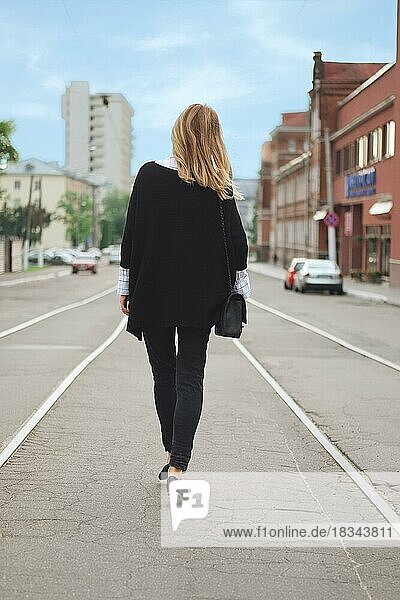 Back view of hipster girl walking on the centre of the street in black poncho and jeans