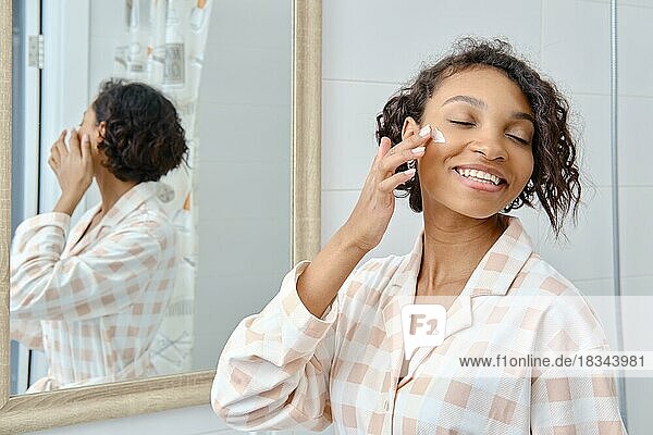 Glückliche afroamerikanische Frau  die Feuchtigkeitscreme auf ihr Gesicht in der Nähe des Spiegels aufträgt
