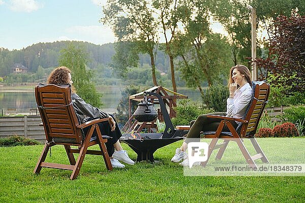 Two women are relaxing in the backyard  sitting in chairs and chatting near the barbecue