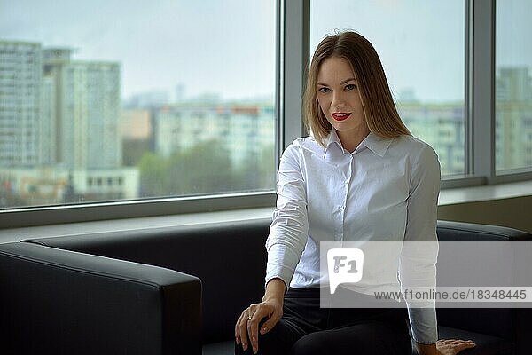 Schöne und positive Geschäftsfrau auf der Couch schaut gerade und lächelnd  Fenster mit Stadtlandschaft im Hintergrund