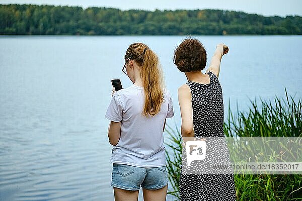 Die Mutter zeigt ihrer Tochter etwas in der Ferne am Ufer des Sees und die Tochter zeichnet eine Route auf dem Smartphone auf