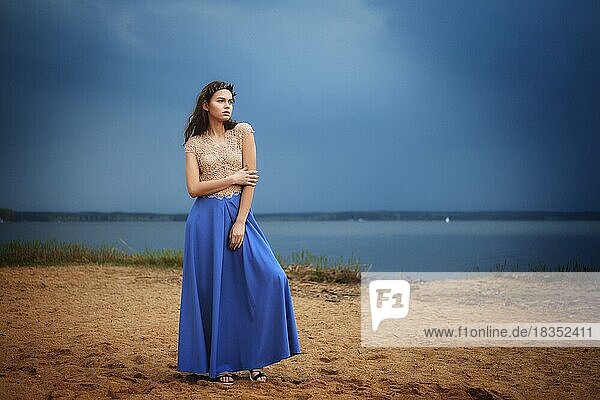 Ruhige einsame Mode-Modell in langen blauen Rock und Spitze Bluse stehend auf einem Strand in einem bewölkten Tag