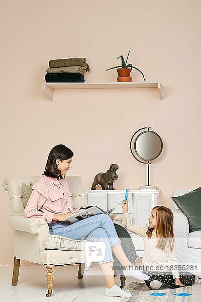 Mutter sitzt im Sessel mit einem Buch und sieht ihrer Tochter zu  die im Wohnzimmer auf dem Boden spielt