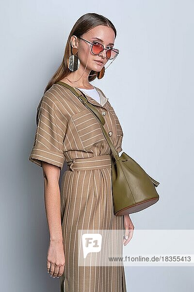 Trendy Dame in großen Sonnenbrille tragen gestreiften olivfarbenen Kleid mit Lederhandtasche posiert über grauen Hintergrund