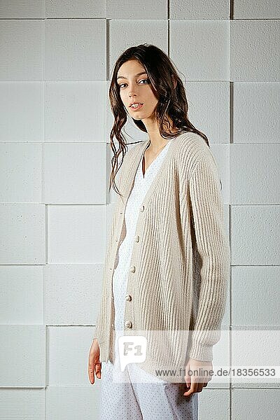 Attractive girl in long knitted woolen sweater posing for look book near gray wall with squares