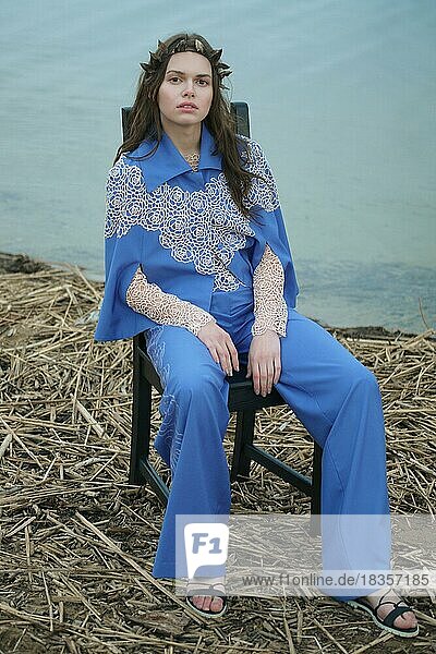 Ruhige einsame Mode-Modell in blauen Hosen und Umhang sitzen auf einem Stuhl in einem bewölkten Tag am Strand