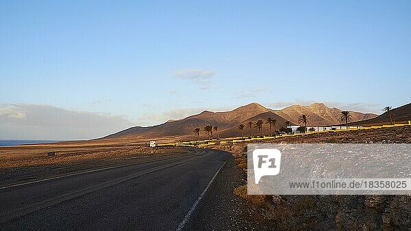 Morgenlicht  Wilder Süden  Jandia  karge Landschaft  nicht-asphaltierte Straße  rotbraune Hügel  Häuser  Palmen  blauer Himmel  wenige Wolken  Fuerteventura  Kanarische Inseln  Spanien  Europa