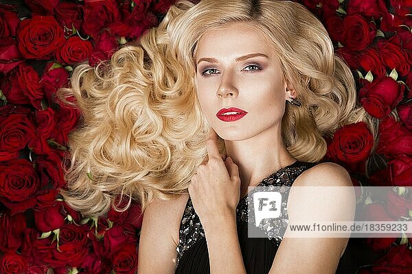Schönes blondes Mädchen auf einem Hintergrund von Rosen liegend. Locken  roter Lippenstift  Abendkleid. Die Schönheit des Gesichts. Fotos im Studio aufgenommen