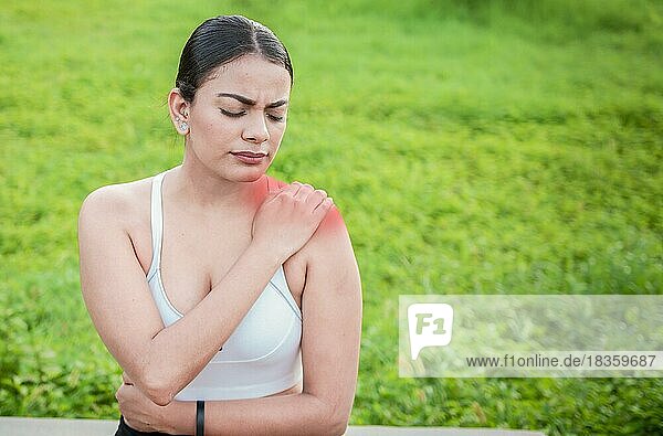 Young woman with shoulder pain sitting outside. Girl suffering from shoulder pain sitting outdoors. Woman sitting and stressed with shoulder pain outdoors. Girl with shoulder pain outdoors