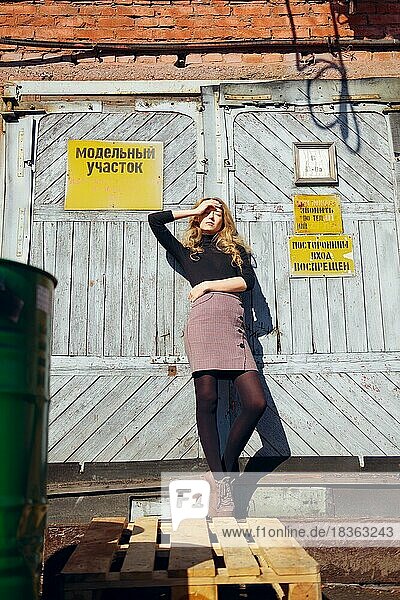 Attraktives Mädchen steht in der Nähe der Fabriktür mit Warnschildern und bedeckt die Augen mit der Hand. (die Aufschrift auf dem Schild ist auf Russisch und kann mit Modellbereich und nicht betreten übersetzt werden)