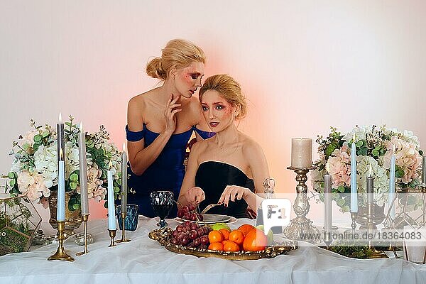 Two fashion models behind the table with fruits  candles and wine. Gossip talks