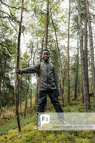 Portrait of smiling boy with stick standing in forest during vacation