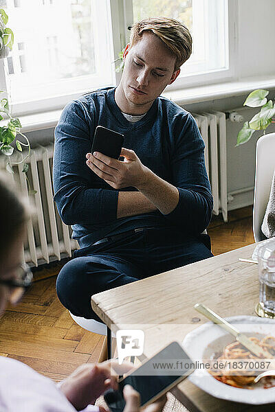 Junger Mann mit Smartphone am Esstisch sitzend in einem Haus
