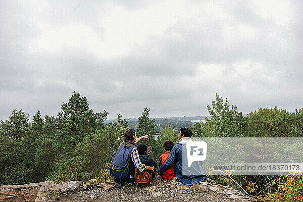 Rear view of woman showing trees to family while sitting on cliff against sky