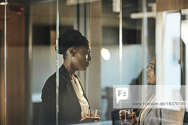 Seitenansicht von Geschäftsfrauen  die im Büro miteinander diskutieren  gesehen durch Glas