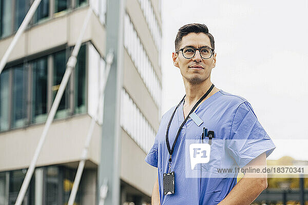 Selbstbewusster männlicher Arzt in blauem Kittel  der vor einem Krankenhausgebäude wegschaut