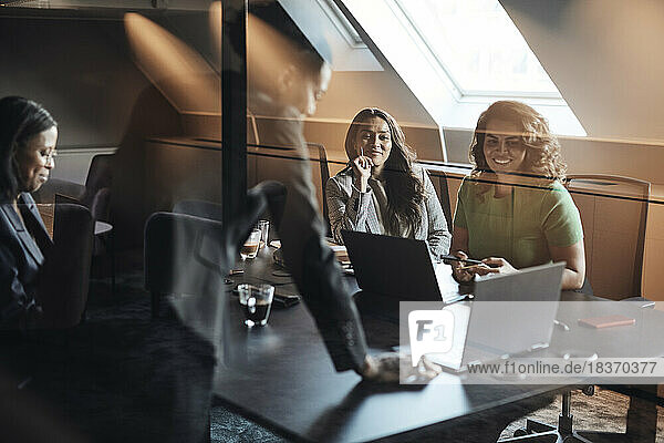 Unternehmerinnen diskutieren über einen Laptop  den sie bei einer Sitzung im Büro durch Glas sehen