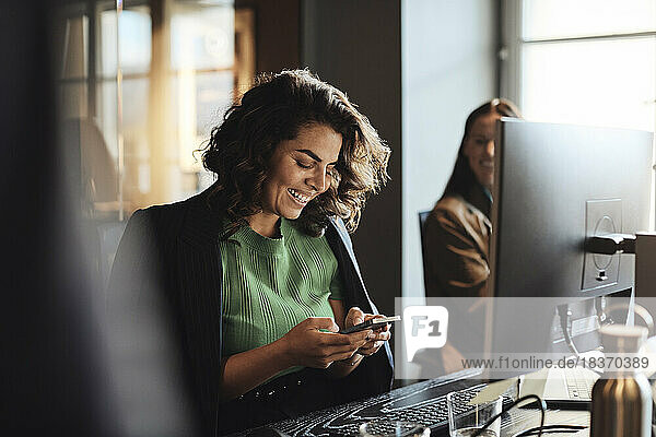 Glückliche Unternehmerin  die am Schreibtisch sitzend im Büro ein Mobiltelefon benutzt