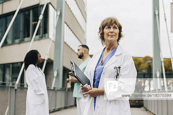 Reife Ärztin schaut weg  während sie ein Klemmbrett und ein Buch außerhalb des Krankenhauses hält