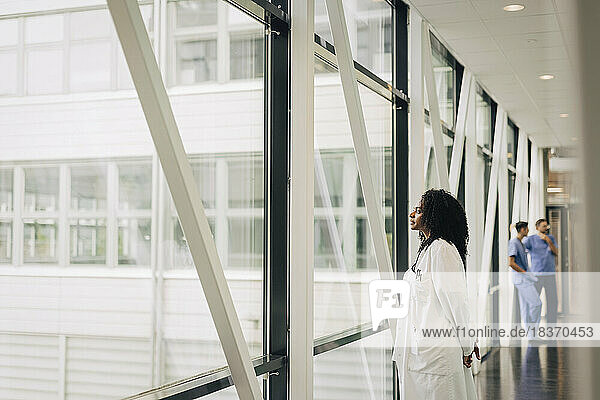 Seitenansicht einer Ärztin  die durch ein Fenster blickt  während sie im Flur eines Krankenhauses steht