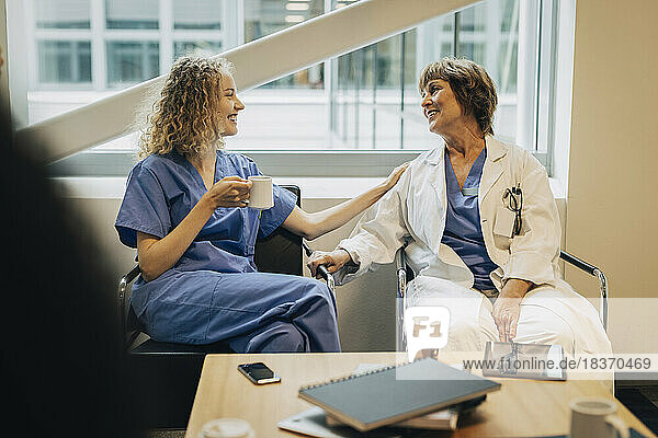 Lächelnde junge Krankenschwester im Gespräch mit einem reifen Arzt  der während der Kaffeepause auf einem Stuhl sitzt