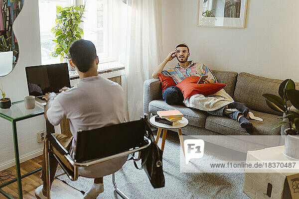 Man on sofa talking to boyfriend sitting with laptop at home