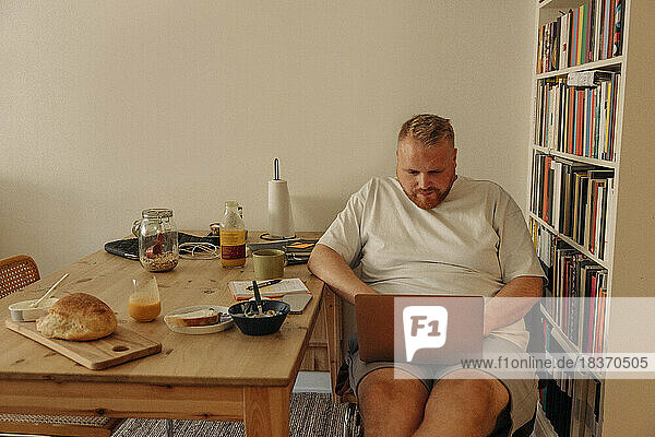 Man using laptop while sitting with breakfast by bookshelf at home