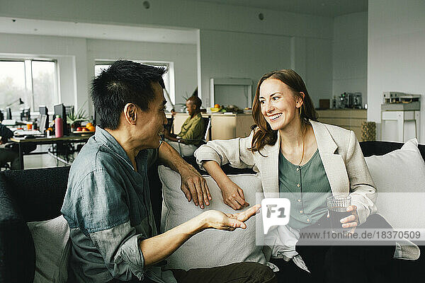 Happy female colleague holding drink while talking to businessman sitting on sofa at home
