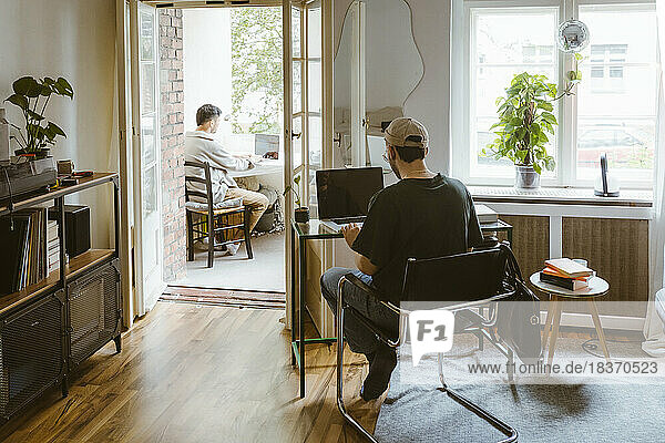 Rear view of gay couple using laptops while sitting on chairs at home