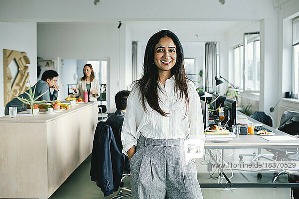 Porträt einer zuversichtlich lächelnden Geschäftsfrau  die mit den Händen in den Taschen im Büro steht
