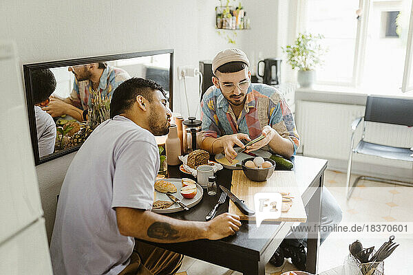 Gay couple sharing smart phone with each other while having food on dining table at home