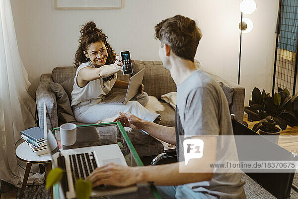 Happy girlfriend showing smart phone to boyfriend with laptop at home