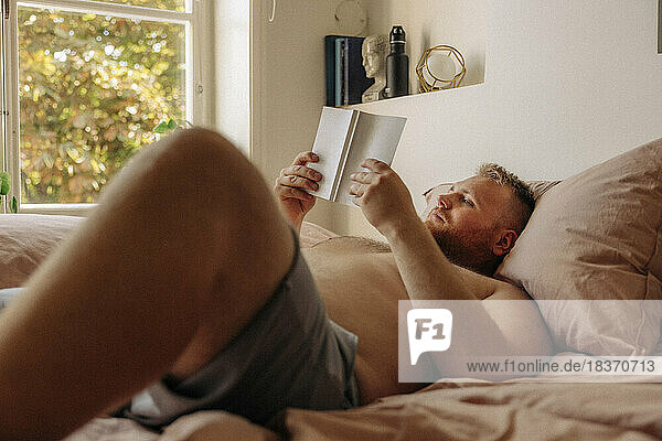 Hemdloser übergewichtiger Mann liest ein Buch  während er zu Hause auf dem Bett liegt