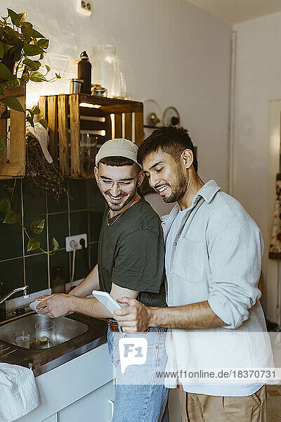 Lächelnder Mann wäscht Geschirr  während sein Freund sein Smartphone in der Küche zu Hause benutzt
