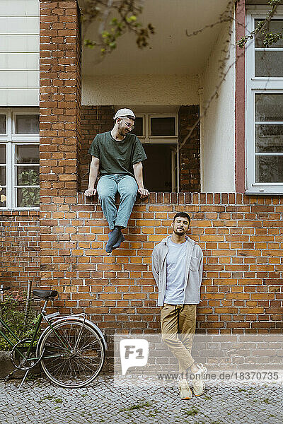 Man looking at boyfriend leaning on brick wall by bicycle outside house
