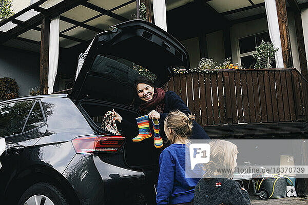 Glückliche Mutter mit Töchtern  die Stiefel und Tasche in den Kofferraum eines Elektroautos laden